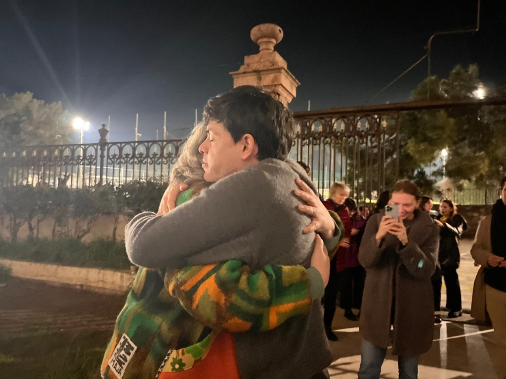 Un hombre con un jersey gris abraza con cariño a una mujer mayor delante de un edificio de aspecto oficial.