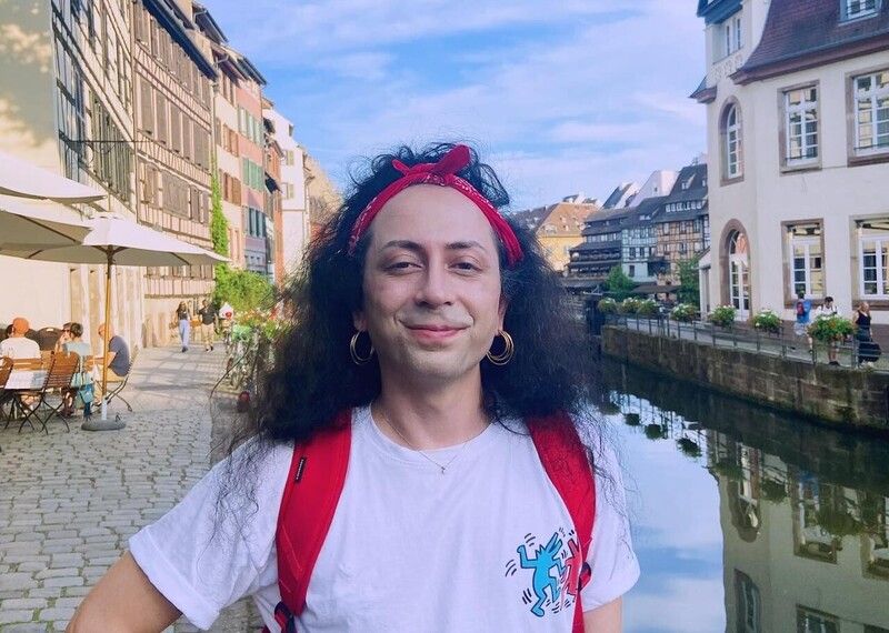 Una persona con una mochila roja y una cinta roja en la cabeza sonríe ampliamente a la cámara junto a un hermoso canal, en cuyas dos orillas hay gente sentada tomando café.