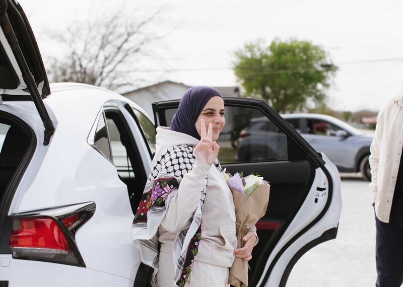 Una mujer vestida con un chándal blanco y un pañuelo negro en la cabeza hace el signo de la paz al salir de un coche blanco.