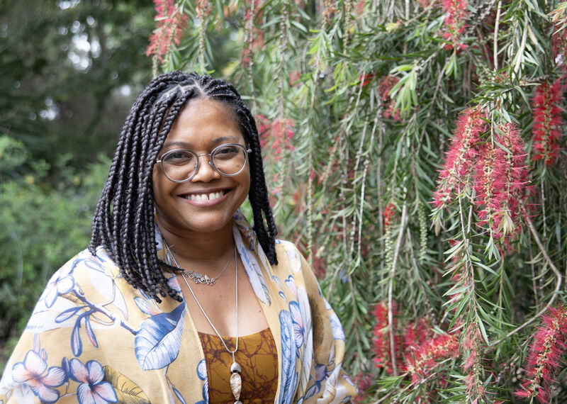 Mujer negra sonriendo a la cámara frente a una árbol florido. Janira Sodré, profesora y educadora comunitaria en Brazil